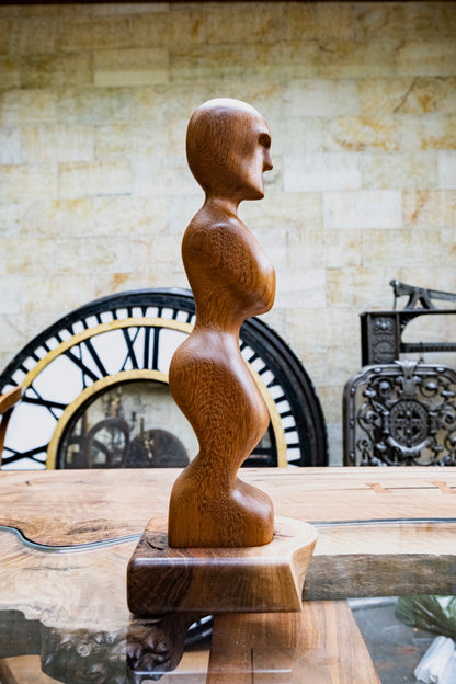 Discover the Artistry of 'Iroko Prayer' with Our Live Edge Sculpture. This captivating wooden statue is a masterpiece, carefully handcrafted to bring elegance to your home. Crafted from the exquisite Iroko wood, known for its healing properties, this remarkable sculpture features a beautifully hand-carved wooden figurine, gracefully mounted on a sleek black walnut raw edge plinth. It's a genuinely unique wood sculpture that infuses any space with timeless beauty.